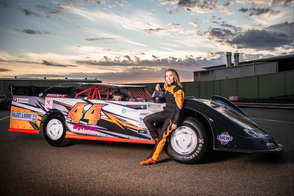 a young woman in an orange and black race driver outfit leans against an orange and black car number 44