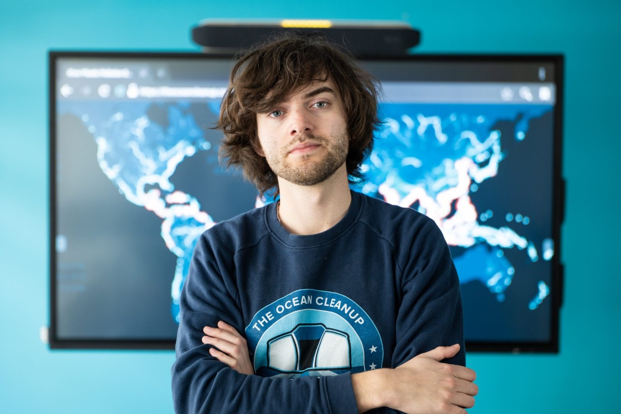 Boyan Slat stands in front of a map of the world in a blue shirt that says 'the ocean cleanup'