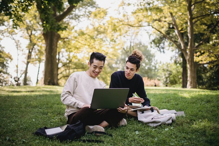 Two young students sit with a laptop outside under trees
